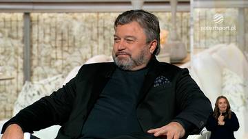 Mężczyzna z siwiejącymi włosami i brodą, ubrany w czarny garnitur i czarną koszulę, siedzi na krześle i mówi.