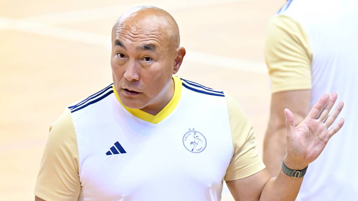 Starszy mężczyzna, prawdopodobnie trener, w białej koszulce z żółtymi elementami i logo, patrzy w bok z lekko otwartymi ustami i uniesioną dłonią.