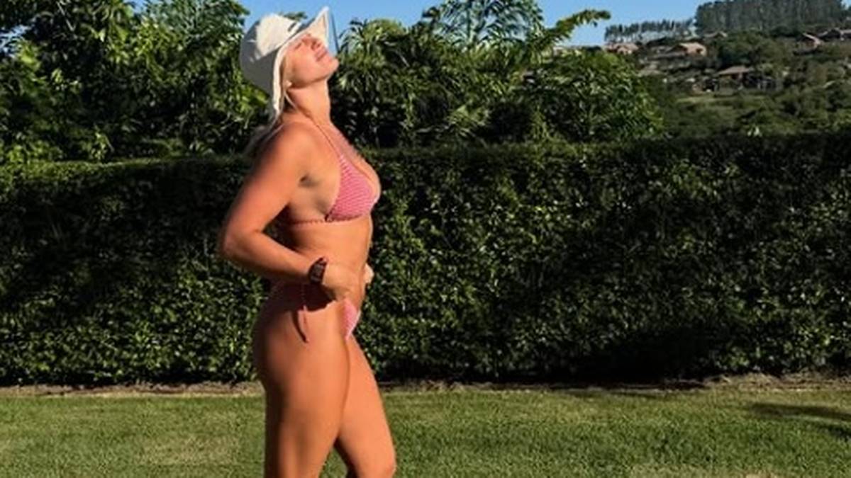 Kobieta w różowym bikini i białym kapeluszu stoi na trawie, z głową uniesioną do słońca. Za nią widać zielony żywopłot i wzgórza.