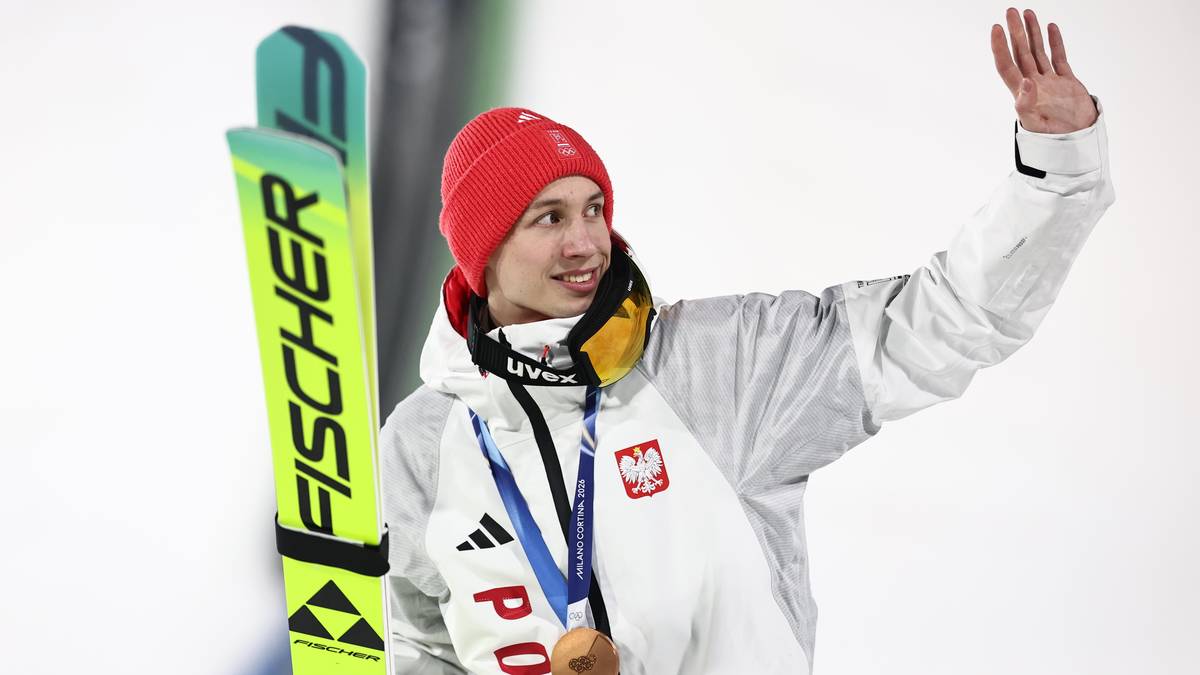 Młody mężczyzna w stroju narciarskim z medalem na szyi i nartami obok, machający do publiczności.