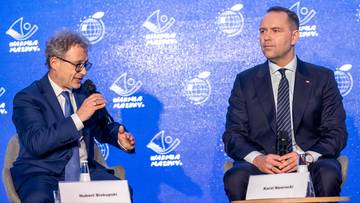 Dw&oacute;ch mężczyzn w garniturach siedzi na krzesłach przed niebieskim tłem z białymi logotypami.