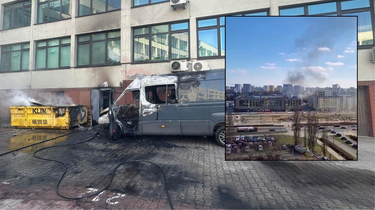 Spalony bus przed budynkiem, z kt&oacute;rego wydobywa się dym. W tle widok miasta z unoszącym się dymem.