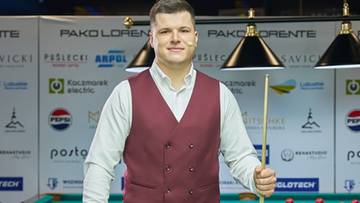 Polski zawodnik bilardowy w kamizelce i koszuli, trzymający kij bilardowy, stoi przy stole bilardowym.