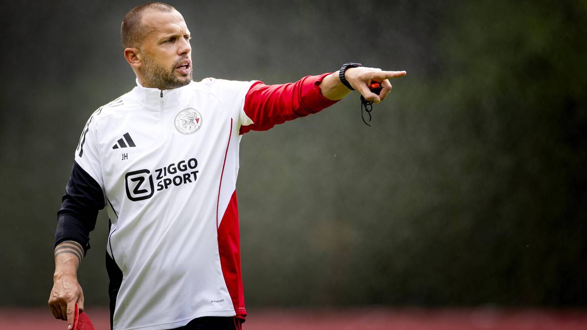 Trener piłkarski Ajaxu w białej koszulce z czerwonymi rękawami wskazuje palcem w prawą stronę.