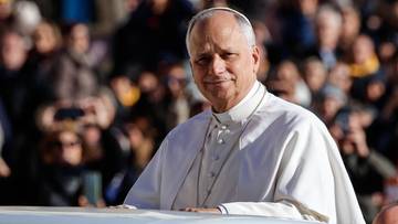 Portret kardynała Leona XIV w białej sutannie, stojącego za białą barierką, z tłumem ludzi w tle.