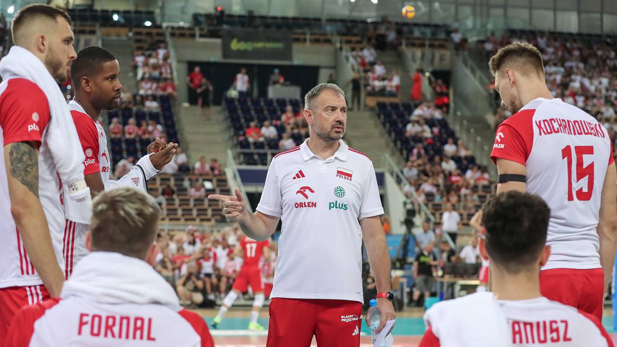 Trener siatk&oacute;wki zwraca się do swoich zawodnik&oacute;w w białych strojach z czerwonymi akcentami, stojąc na boisku w otoczeniu publiczności na trybunach.