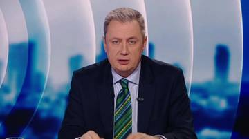 Mężczyzna w ciemnym garniturze i pasiastym krawacie siedzi za stołem, patrząc wprost.