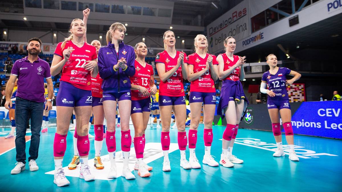 Drużyna siatkarek w strojach sportowych, w tym czerwonych koszulkach z numerami i niebieskich spodenkach, stoi na boisku do siatk&oacute;wki, niekt&oacute;re klaszczą. Obok nich stoi trener w fioletowej koszulce. W tle widać trybuny i tablice wynik&oacute;w.