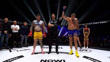 Dwóch zawodników stoi na ringu bokserskim, jeden z uniesioną ręką jako zwycięzca, w tle widać sędziów i publiczność.