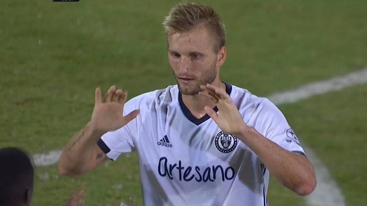 Piłkarz w białej koszulce z widocznym logo klubu i napisem "Artesano" na zielonej murawie boiska.