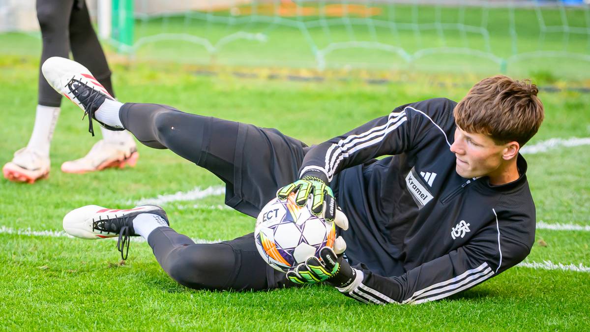 Piłkarz w stroju bramkarskim leży na trawie i łapie piłkę.