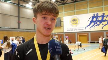 Młody mężczyzna z medalem na szyi udziela wywiadu przed mikrofonem w hali sportowej.