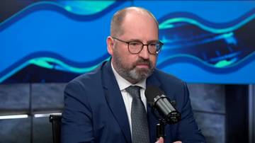 Mężczyzna w okularach i granatowym garniturze siedzi przy mikrofonie, patrząc w d&oacute;ł.