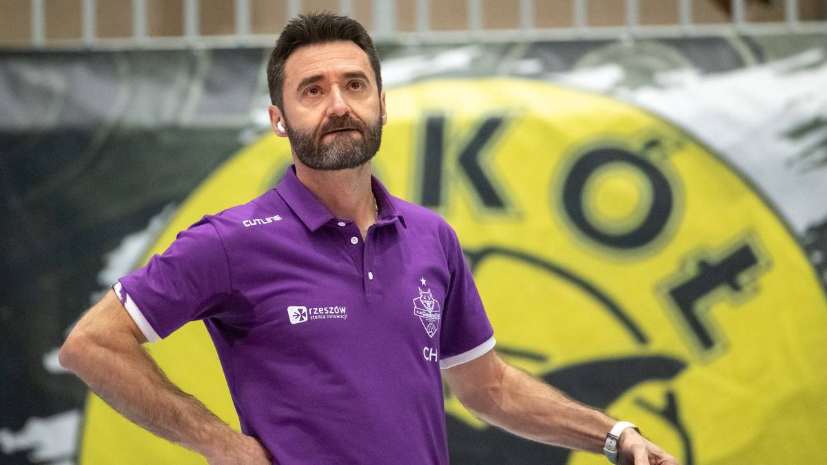 Mężczyzna w fioletowej koszulce stoi na tle baneru z logo klubu.