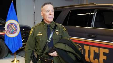 Funkcjonariusz w mundurze stoi obok niebieskiej flagi ze złotym emblematem i samochodu z napisem ICE.