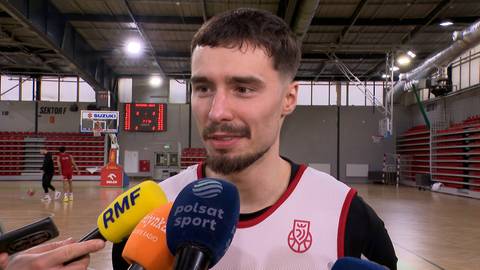 Mężczyzna w stroju sportowym z mikrofonami skierowanymi w jego stronę, udzielający wywiadu na sali gimnastycznej.