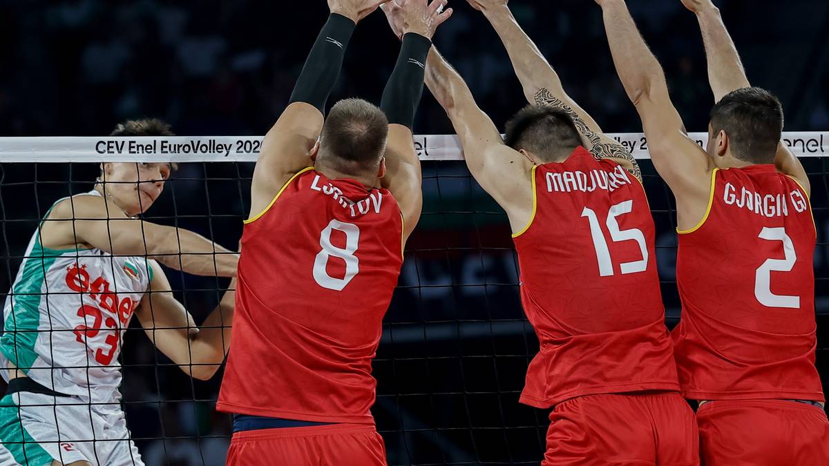 Czterech siatkarzy blisko siatki podczas meczu. Trzech graczy w czerwonych koszulkach w akcji blokowania, a jeden gracz w białej koszulce z numerem 33 odbija piłkę.