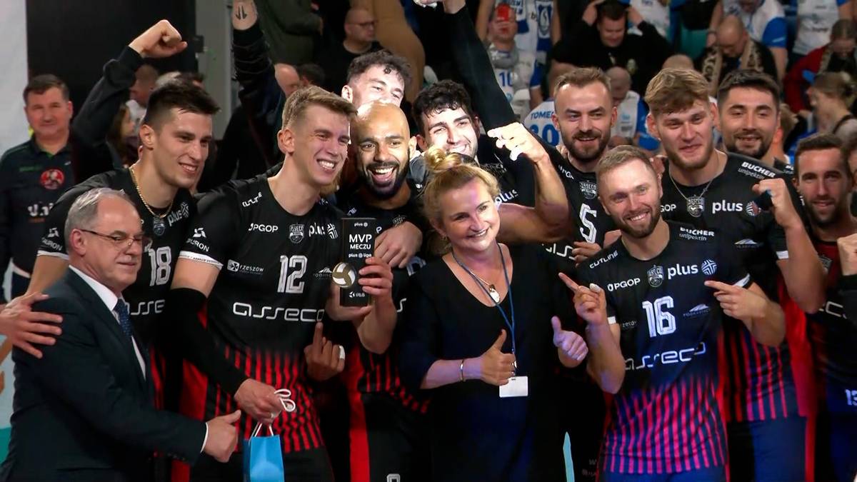 Drużyna siatkarzy w czarnych strojach z czerwonymi elementami, świętująca zwycięstwo. Na pierwszym planie stoi trener, otoczony przez zawodników trzymających puchar.