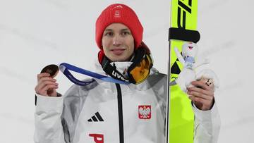 Polski skoczek narciarski w stroju reprezentacyjnym trzyma medal olimpijski i narty.