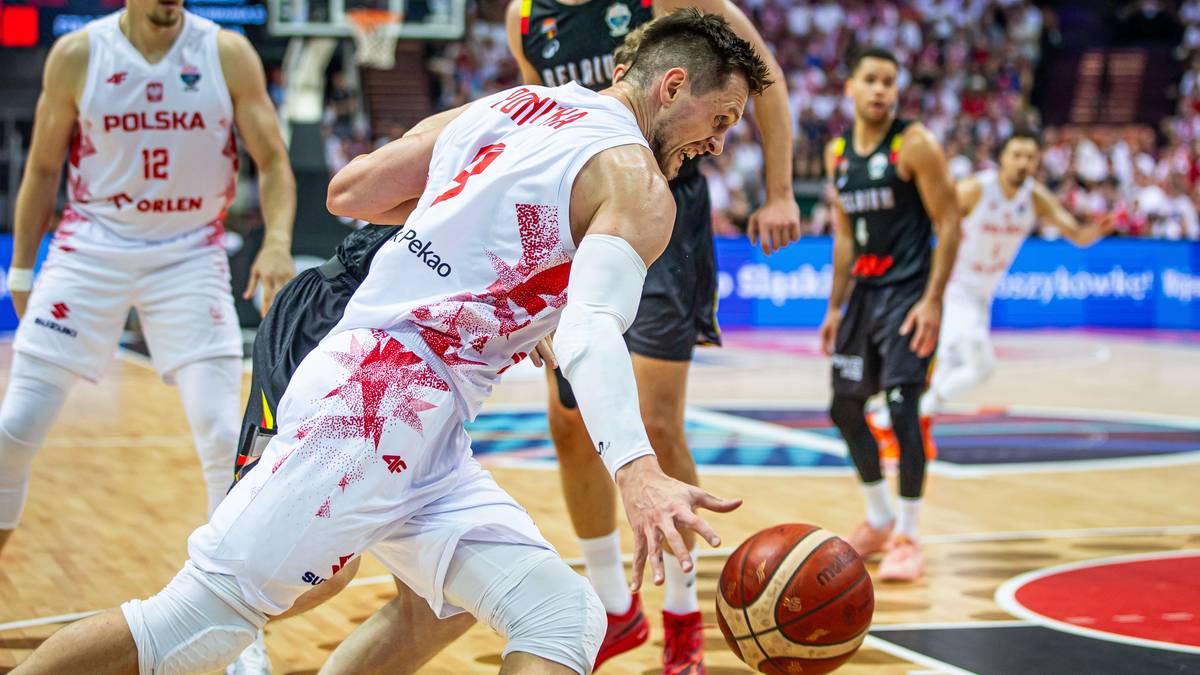 Koszykarz w białym stroju z czerwonymi zdobieniami pochyla się nad piłką do koszykówki na boisku.