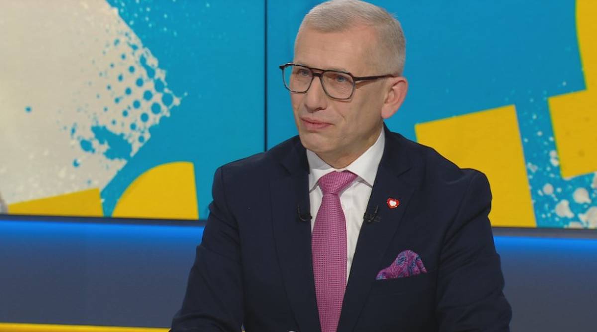 Mężczyzna w garniturze i okularach siedzi i patrzy w bok.