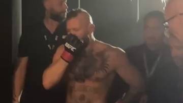 Conor McGregor w rękawicach bokserskich, z ręką na twarzy, otoczony przez innych ludzi.