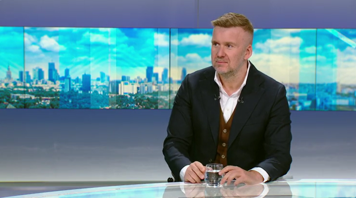 Mężczyzna w garniturze siedzi przy stole i patrzy w bok. Za nim znajduje się ekran z widokiem na panoramę miasta.