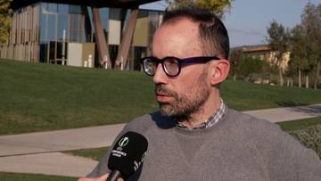 Mężczyzna w okularach udzielający wywiadu przed nowoczesnym budynkiem sportowym.