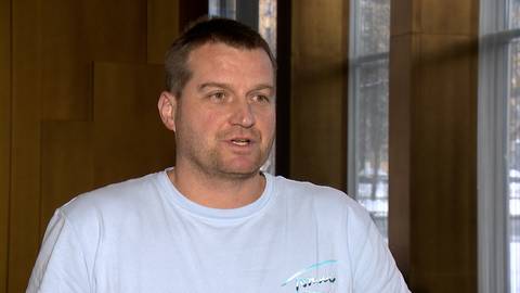 Mężczyzna z kr&oacute;tkimi włosami i zarostem, ubrany w jasnoniebieską koszulkę, wypowiada się na tle okien i drewnianych paneli.