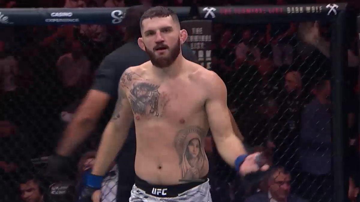 Biały mężczyzna w rękawicach bokserskich, w białym stroju sportowym z logo UFC, stoi w klatce oktagonu, patrząc przed siebie.