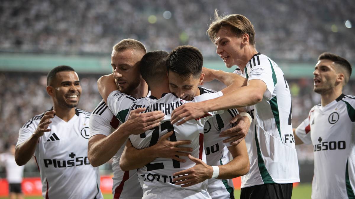 Pięciu piłkarzy w białych strojach Legii Warszawa obejmuje się w radosnym uścisku na murawie stadionu.