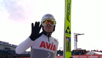 Skoczek narciarski w kombinezonie i kasku z goglami, machający ręką, obok niego stoi narta.