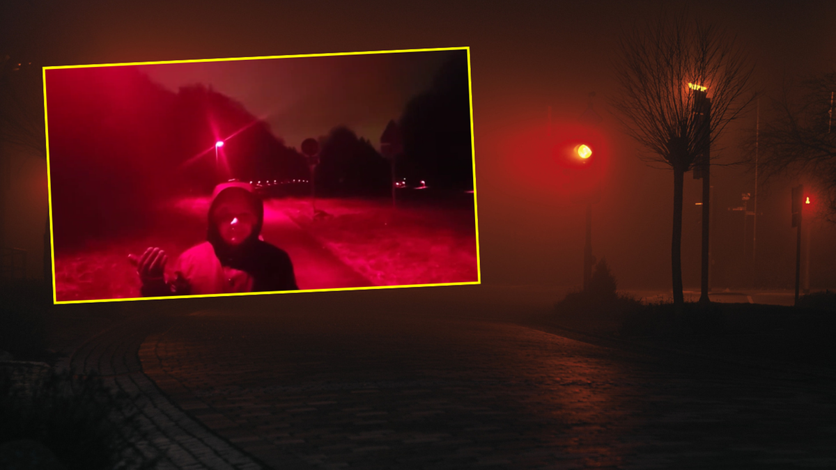 Młoda osoba w kapturze stojąca na drodze w nocy, oświetlona czerwonym światłem z latarni ulicznej.