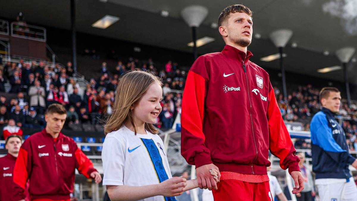 Piłkarz reprezentacji Polski w czerwonej kurtce sportowej idzie obok dziewczynki w białej koszulce piłkarskiej, trzymając ją za rękę. W tle widoczni inni piłkarze oraz trybuny stadionu wypełnione kibicami.