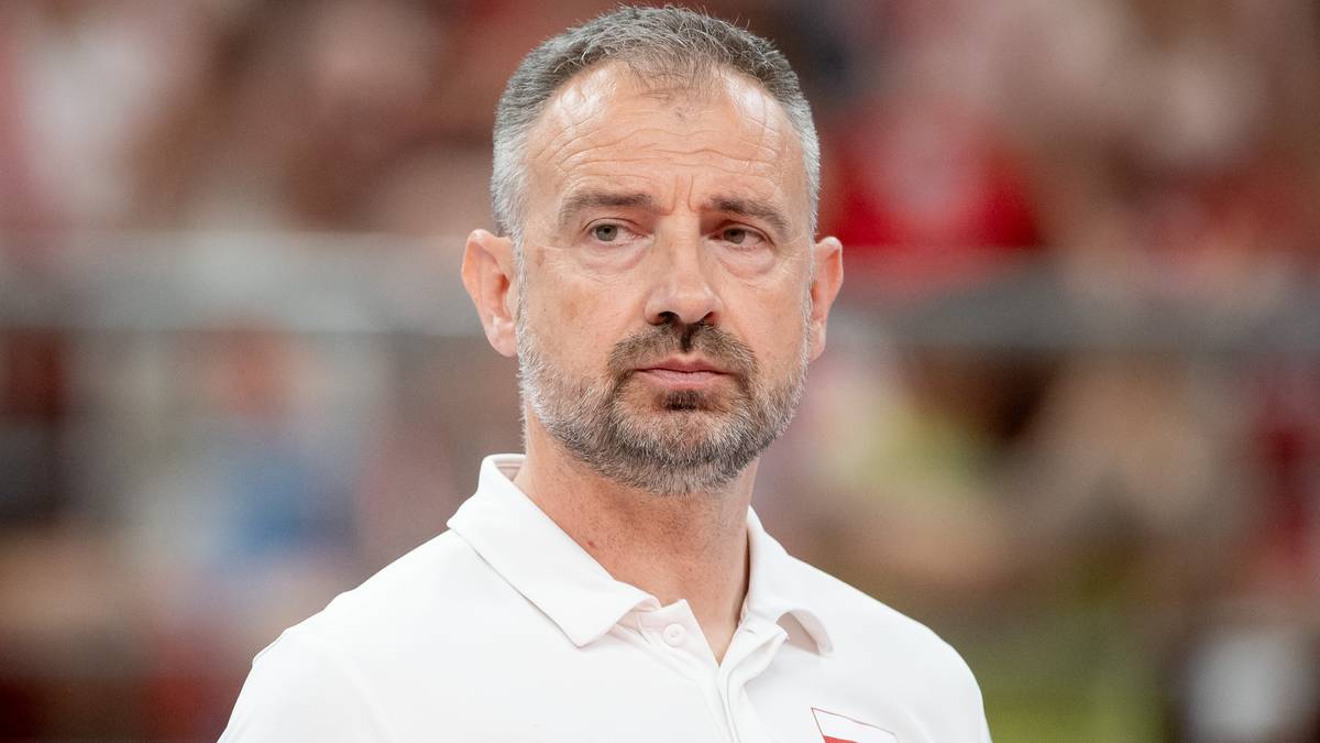 Portret mężczyzny w białej koszulce polo, o krótkich, siwych włosach i zarostem. Wyraz twarzy jest poważny, z lekkim zamyśleniem. Tło jest rozmyte, sugerujące obiekt sportowy.