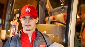 Młody mężczyzna w czerwonej czapce z daszkiem z logo NY i szarej kurtce, patrzący w obiektyw.