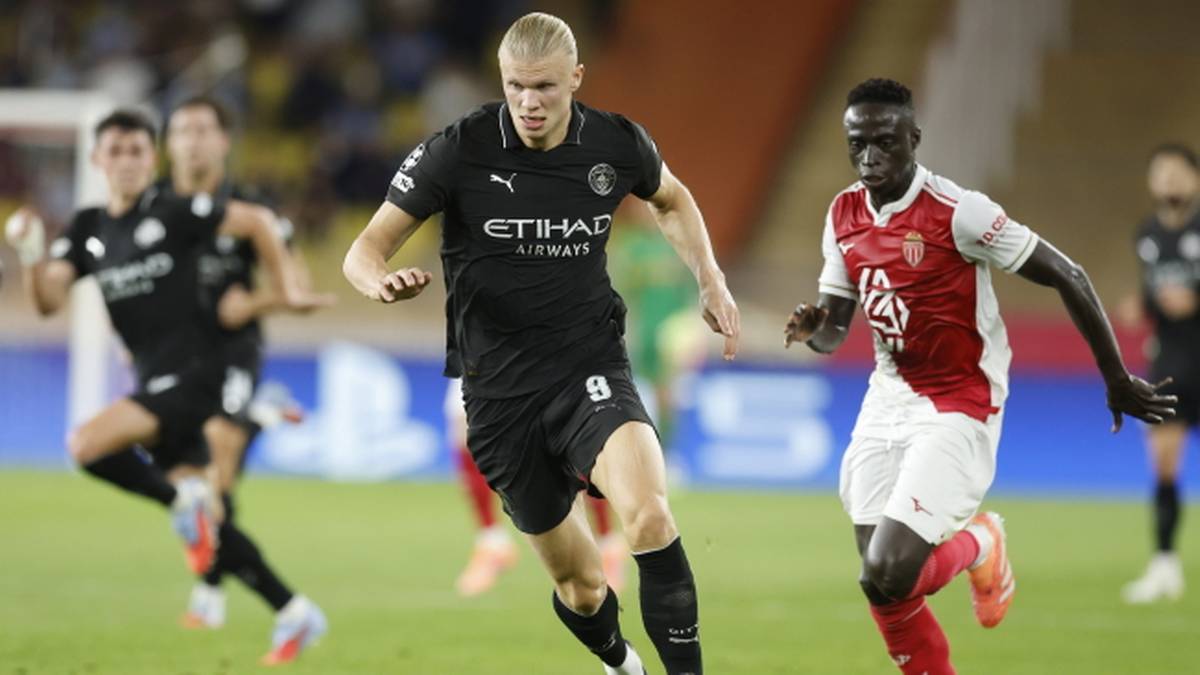 Piłkarz Erling Haaland w czarnej koszulce Manchesteru City, numer 8, biegnie z piłką po zielonej trawie boiska podczas meczu. Na jego tle widać innych zawodników w czerwono-białych strojach oraz trybuny.