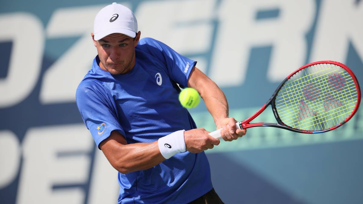 Tenisista w niebieskiej koszulce i białej czapce uderza piłkę rakietą podczas meczu.