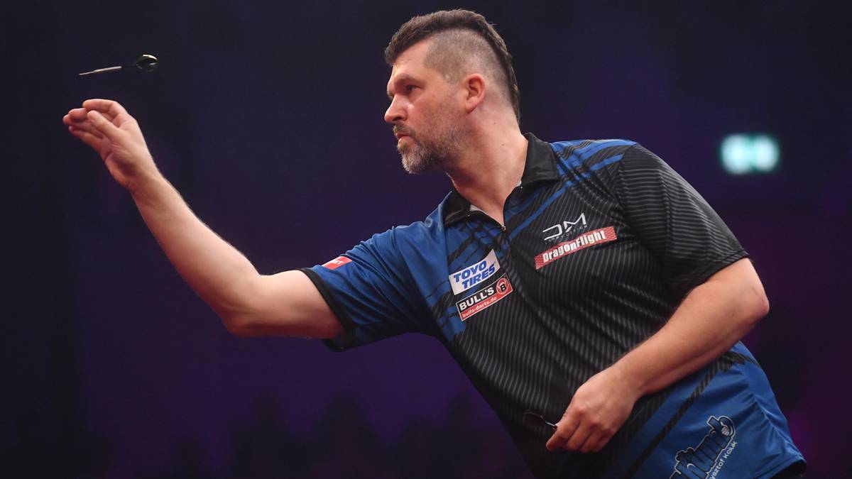 Mężczyzna w ciemnej koszulce z niebieskimi wstawkami rzuca rzutką do celu.