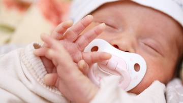 Zbliżenie na twarz śpiącego niemowlęcia z rączkami przed twarzą i smoczkiem w ustach.