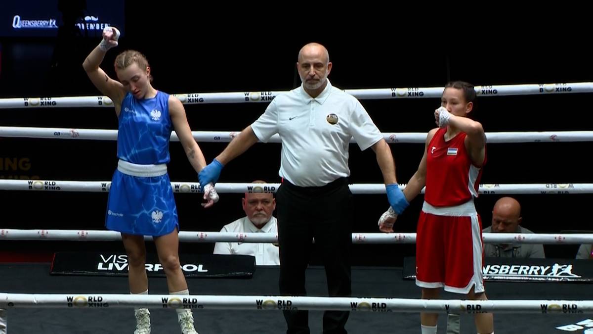 Dwie bokserki w czerwonych i niebieskich strojach stoją na ringu, połączone dłońmi, z sędzią pośrodku.