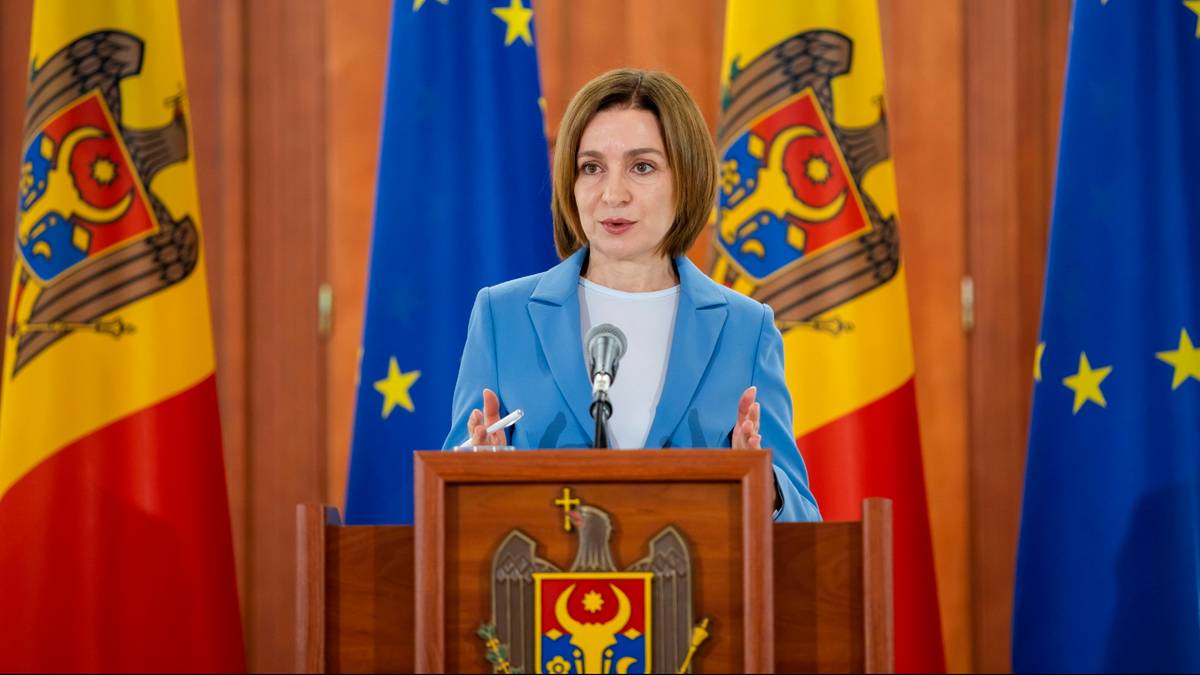 Kobieta w niebieskiej marynarce mówi do mikrofonu, stojąc za podium z herbem. W tle widoczne flagi Mołdawii i Unii Europejskiej.