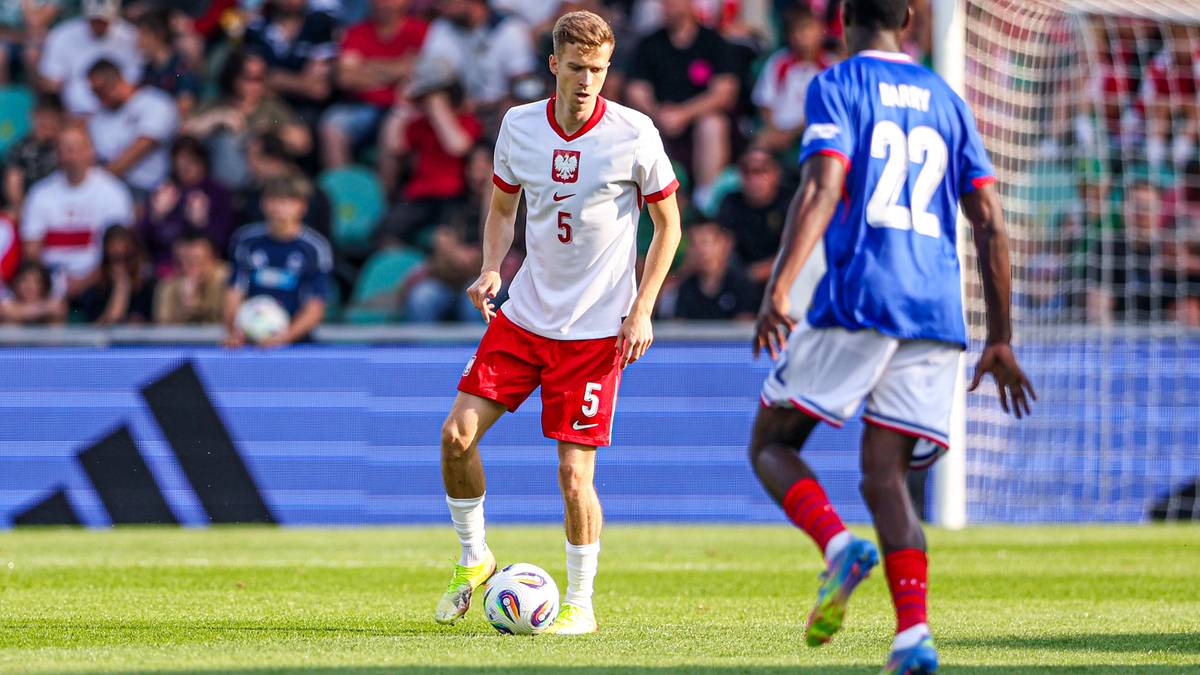 Piłkarz w białej koszulce z numerem 5 i czerwonymi spodenkami prowadzi piłkę, a obok niego stoi zawodnik w niebieskiej koszulce z numerem 22.