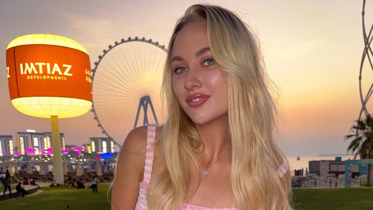 Młoda kobieta z blond włosami i w letniej sukience stoi na tle panoramy miasta o zachodzie słońca. Widoczny jest diabelski młyn.