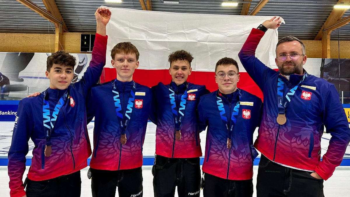 Pięciu młodych mężczyzn w strojach sportowych z medalem na szyi i flagą Polski, stojący na lodowisku do curlingu.