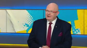 Łysy mężczyzna w okularach i granatowym garniturze z bordowym krawatem i poszetką, mówiący w studio telewizyjnym.