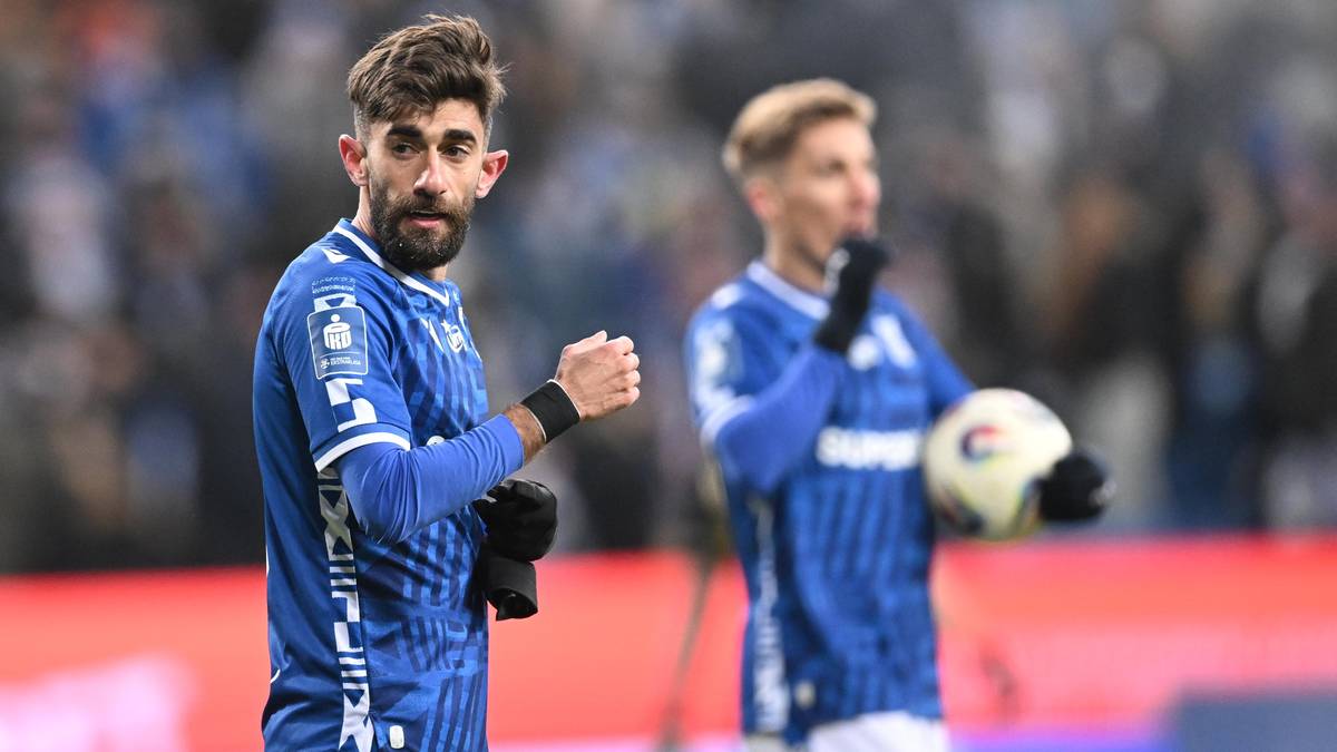 Dw&oacute;ch piłkarzy w niebieskich strojach na boisku piłkarskim, jeden z nich trzyma piłkę.