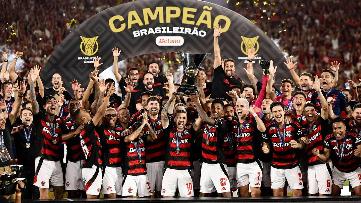 Piłkarze Flamengo Rio de Janeiro świętujący zdobycie ósmego tytułu mistrza Brazylii, trzymający puchar, otoczeni przez kibiców pod łukiem z napisem "CAMPEÃO BRASILEIRÃO"