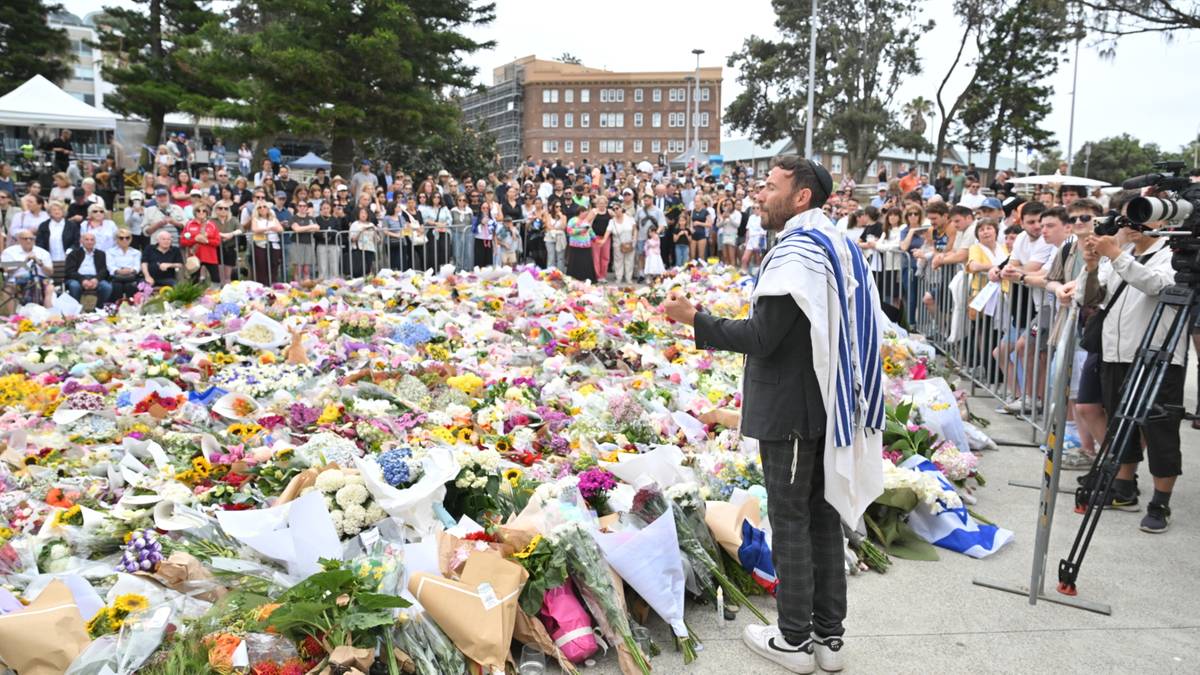 Tłum ludzi zgromadzonych przed ogromną ilością kwiatów złożonych jako hołd, z mężczyzną stojącym przodem do zgromadzonych.