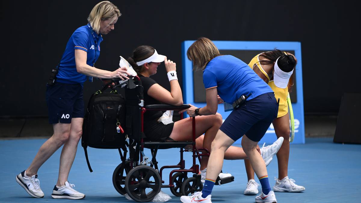 Zawodniczka tenisa siedzi na w&oacute;zku inwalidzkim, otoczona przez trzy inne osoby. Dwie z nich pomagają jej wstać, trzymając ją pod ramiona. Jedna osoba stoi obok, a kolejna klęczy z tyłu. Wszyscy ubrani są w sportowe stroje.